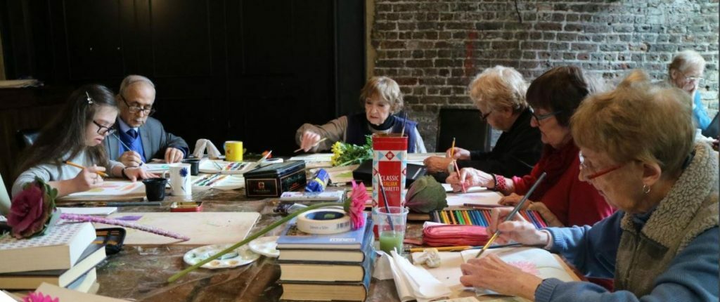 A group of people sitting around a table together. Each person has a box of watercolour paints and they are painting on paper.
