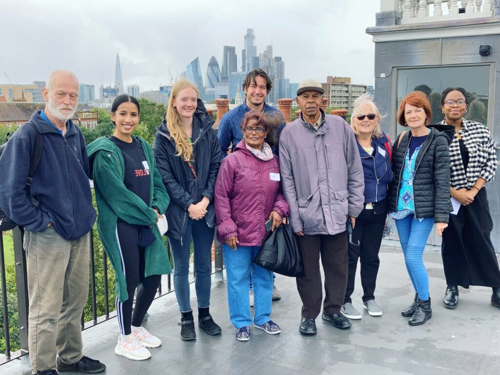 9 people of various ages standing in a group and smiling. Behind them is a view of London.