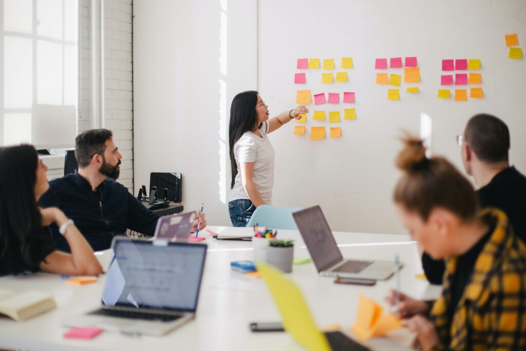 Colleagues sit around a table with their laptops open. One person is standing up and pointing at a white board which has lots of post-it notes on it.