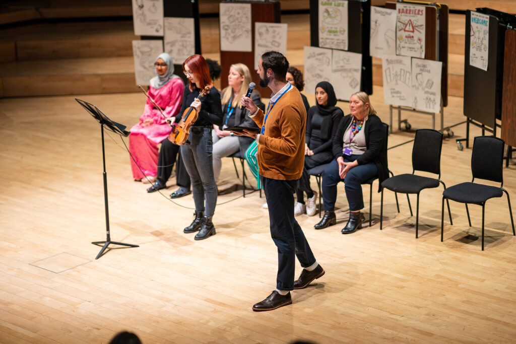 A live performance of Hidden. A group of people perform in front of an audience, one performer holds a violin and another performer has a microphone