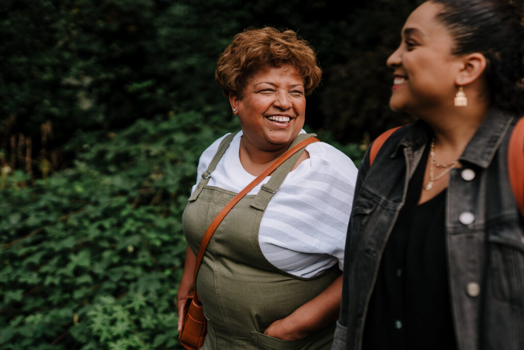 Two women walking and laughing together outside, against a background of leafy green foliage