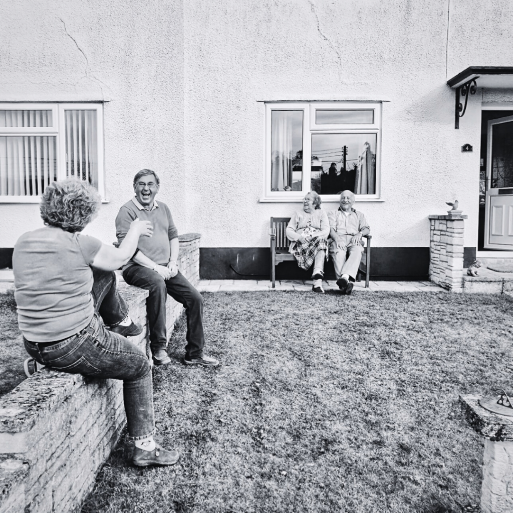 Black and white photo. A group of four older people sitting outside in a garden. 2 of the group are sitting together on a garden bench and smiling. The other 2 are sitting on a small brick wall, facing each other and talking happily