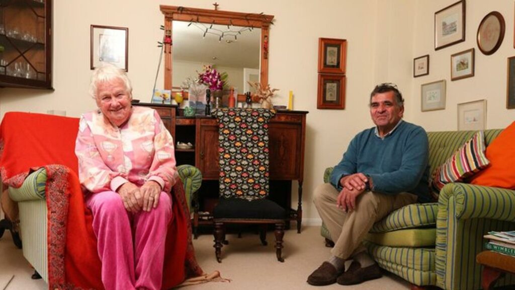 An older woman and an older man sitting on comfy chairs in their living room. They are both smiling at the camera.