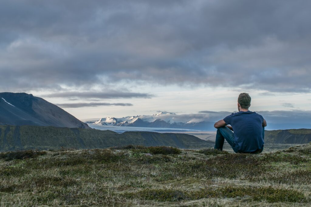 A man sitting down on top of a hill, overlooking a vast landscape of mountains, river and a cloudy sky.