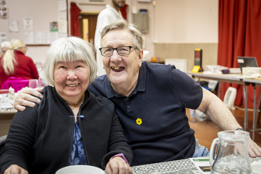 An older man and an older woman smiling widely at the camera. The man has his arm resting on the woman's shoulder