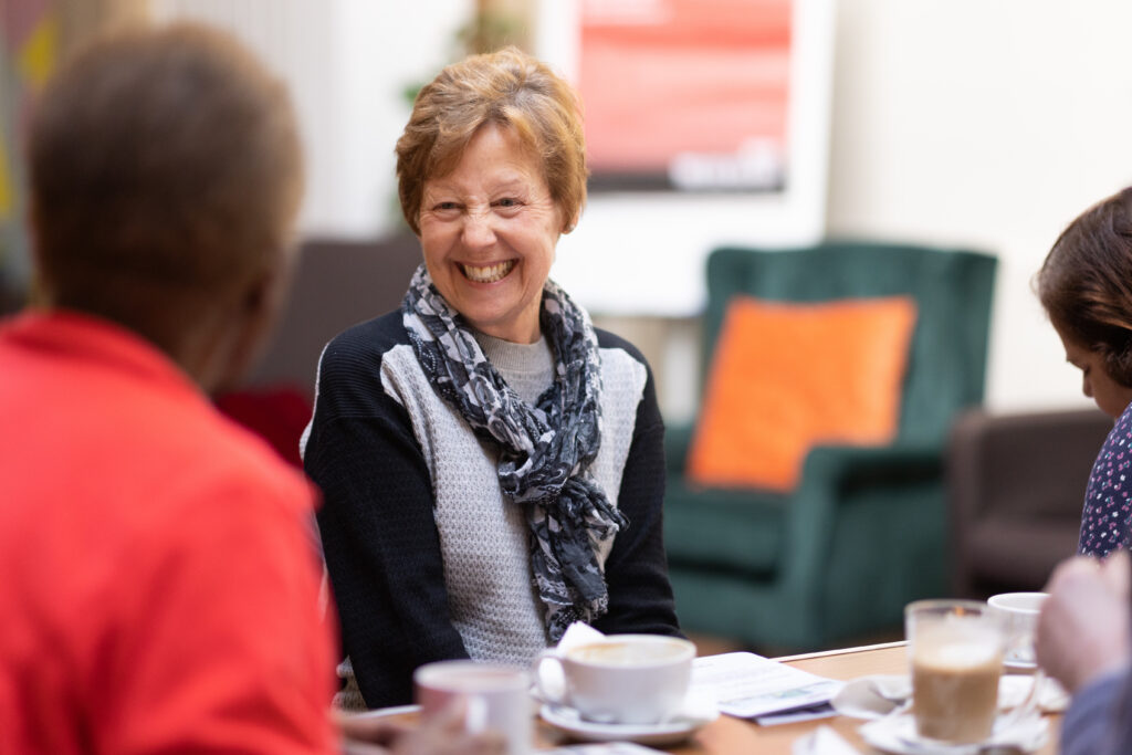 An older women sitting at a table with a cup in front of her. She is smiling widely at someone else who is sitting close to her, of whom we can see the back of their head