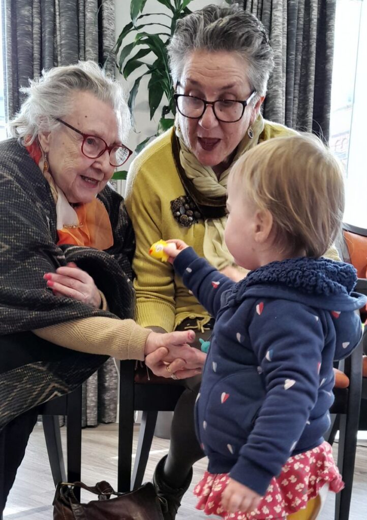 Two older women smiling and holding out their hands to a toddler who is handing them a small yellow toy.