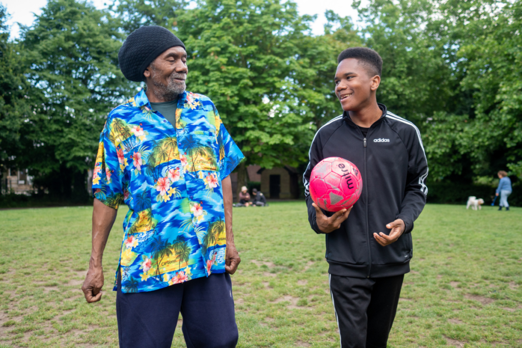 An older person and younger person outside in a park. The younger person is holding a football.