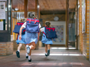 Three young girls with backpacks running down a school hallway.