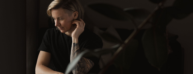 Image of the author - Anna Sobieniak. with short blonde hair, dressed in a black t-shirt, sits quietly in a dimly lit room with a contemplative expression, their hand resting near their face.