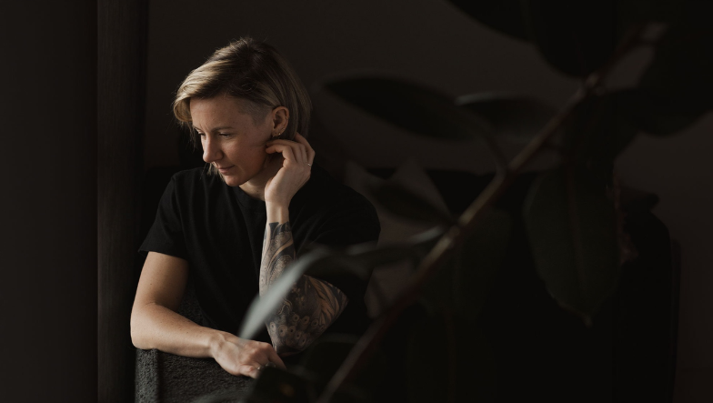 Image of the author - Anna Sobieniak. with short blonde hair, dressed in a black t-shirt, sits quietly in a dimly lit room with a contemplative expression, their hand resting near their face.