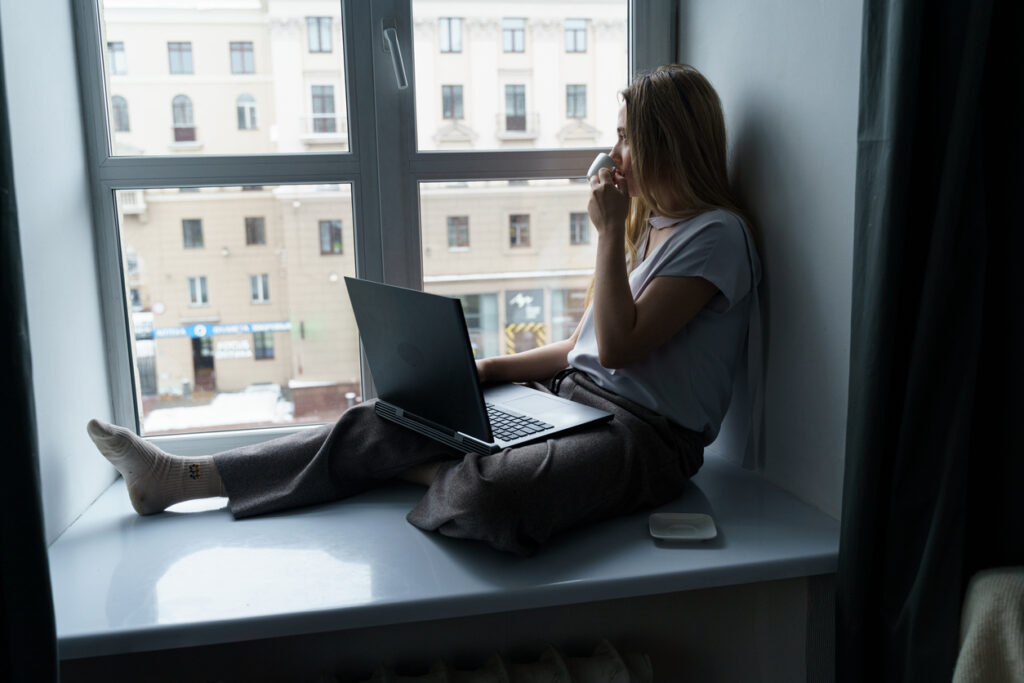 A woman sitting on a windowsill with a laptop on her lap, holding a cup of coffee and gazing out the window in a reflective moment.