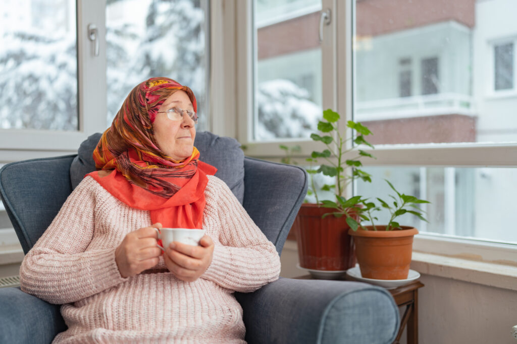 An old muslim woman is sitting on sofa enjoying drinking hot drink and relaxing in the living room at home while it is snowing and cold outside.