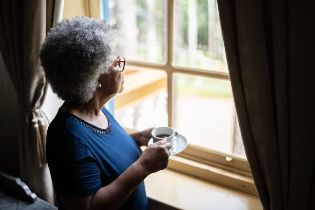 A Contemplative senior woman looks through the window while drinking coffee at home