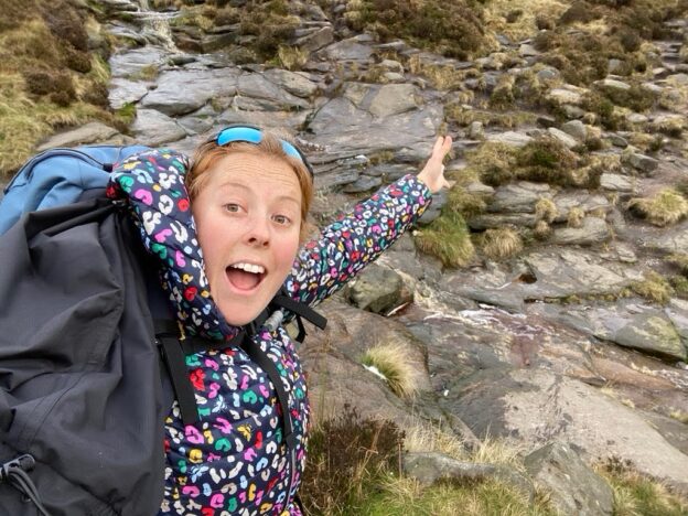 A woman by a stream waving animatedly at the camera