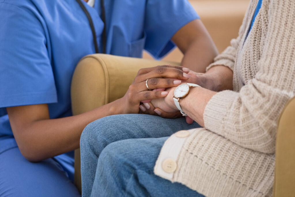 Part of female home caregiver supporting senior woman in her house, holding hands.