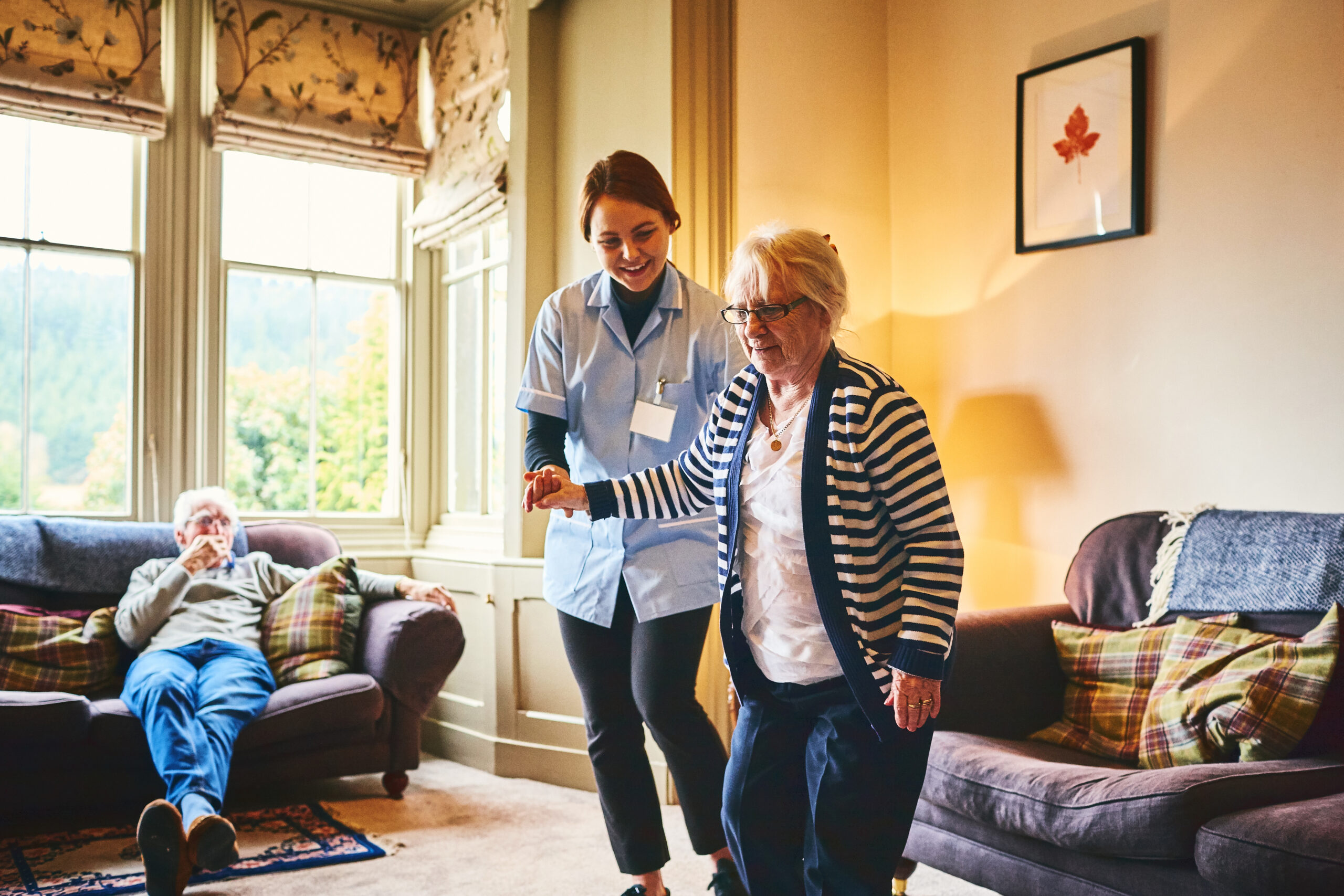 Young female nurse supporting old woman to walk indoors with senior man relaxing on sofa at back