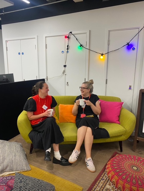 2 attendees sat facing each other on an olive green sofa with pink and orange cushions talking animatedly