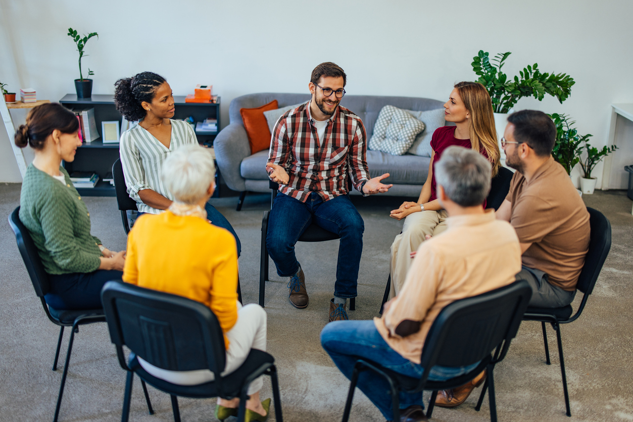 Group of people sitting in a circle talking to each other, having a group therapy appointment.