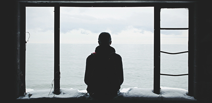 A person sitting on a window ledge looking out at a ocean.