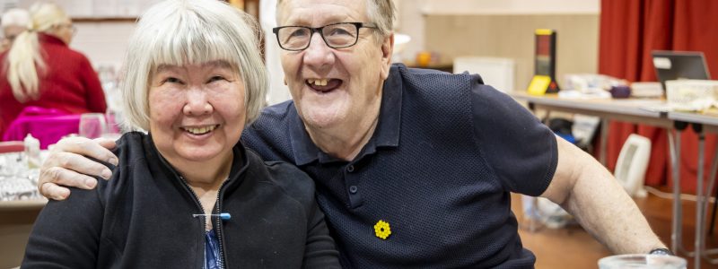 An older man and an older woman smiling widely at the camera. The man has his arm resting on the woman's shoulder