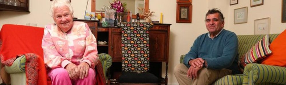 An older woman and an older man sitting on comfy chairs in their living room. They are both smiling at the camera.