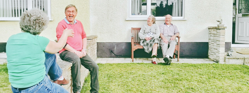 A group of four older people sitting outside in a garden. 2 of the group are sitting together on a garden bench and smiling. The other 2 are sitting on a small brick wall, facing each other and talking happily